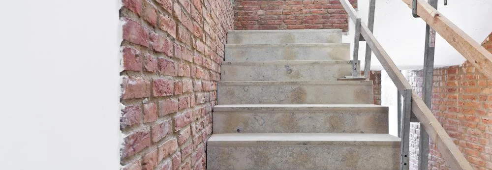 Eine Betontreppe im Rohbau mit einer Backsteinwand und einem provisorischen Geländer.